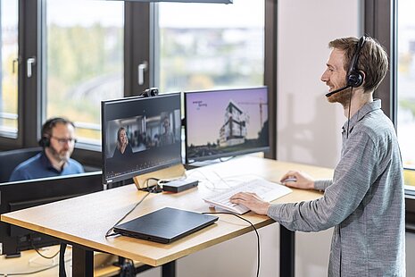 Person mit Headset steht am Schreibtisch und blickt auf zwei Monitore.