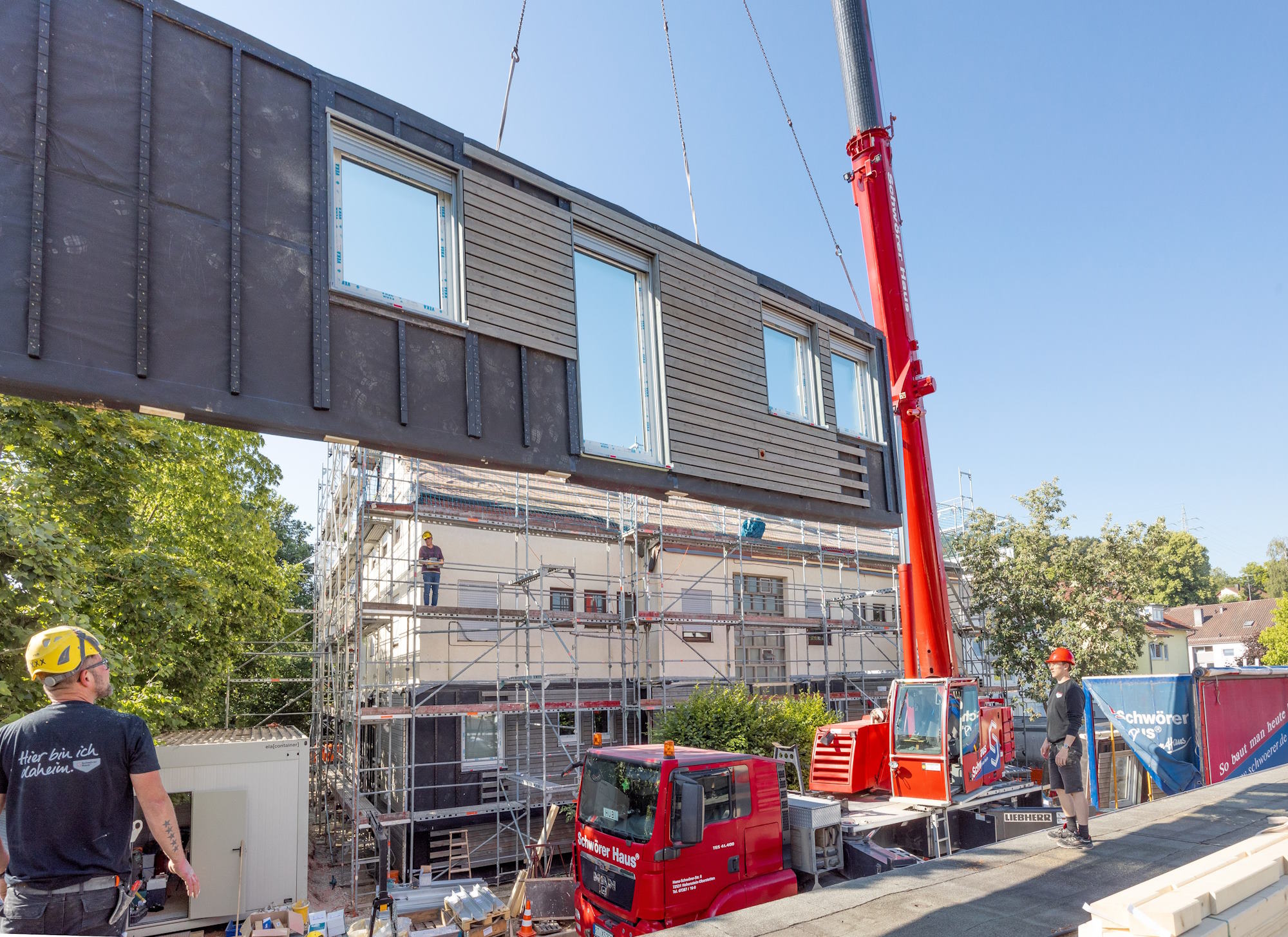 Schwebendes Fassadenelement mit Fenstern an Kranfahrzeug, drei Bauarbeiter schauen stehen und schauen