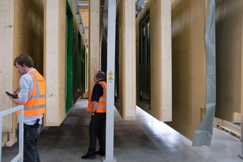 Bild zeigt: hängende Fassadenelemente in Fabrik und zwei Männer in Warnwesten davor