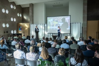 Personen sitzen auf Stühlen in einem Veranstaltungsraum und schauen zur Bühne.