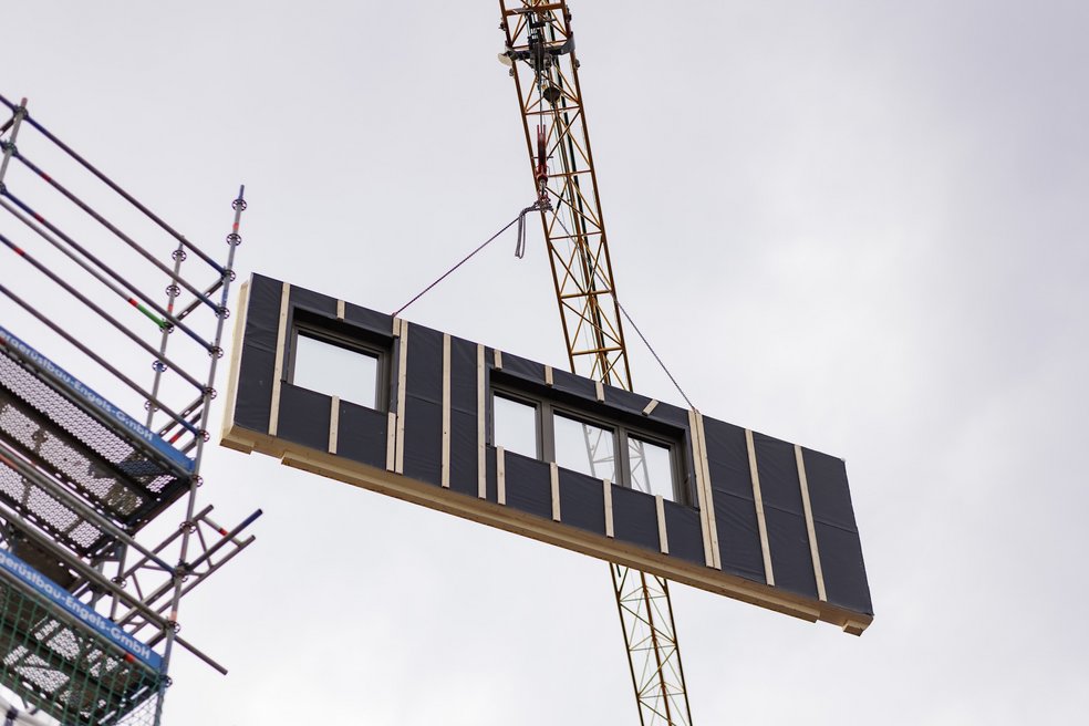 Vorgefertigtes Hausfassadenelement mit Fenstern schwebt an Kran.