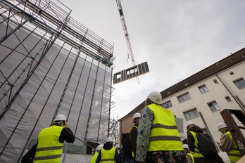 Personen in gelben Sicherheitswesten und mit Bauhelmen auf dem Kopf stehen vor einem Gebäude und schauen nach oben zu einem Fassadenelement an einem Kran.