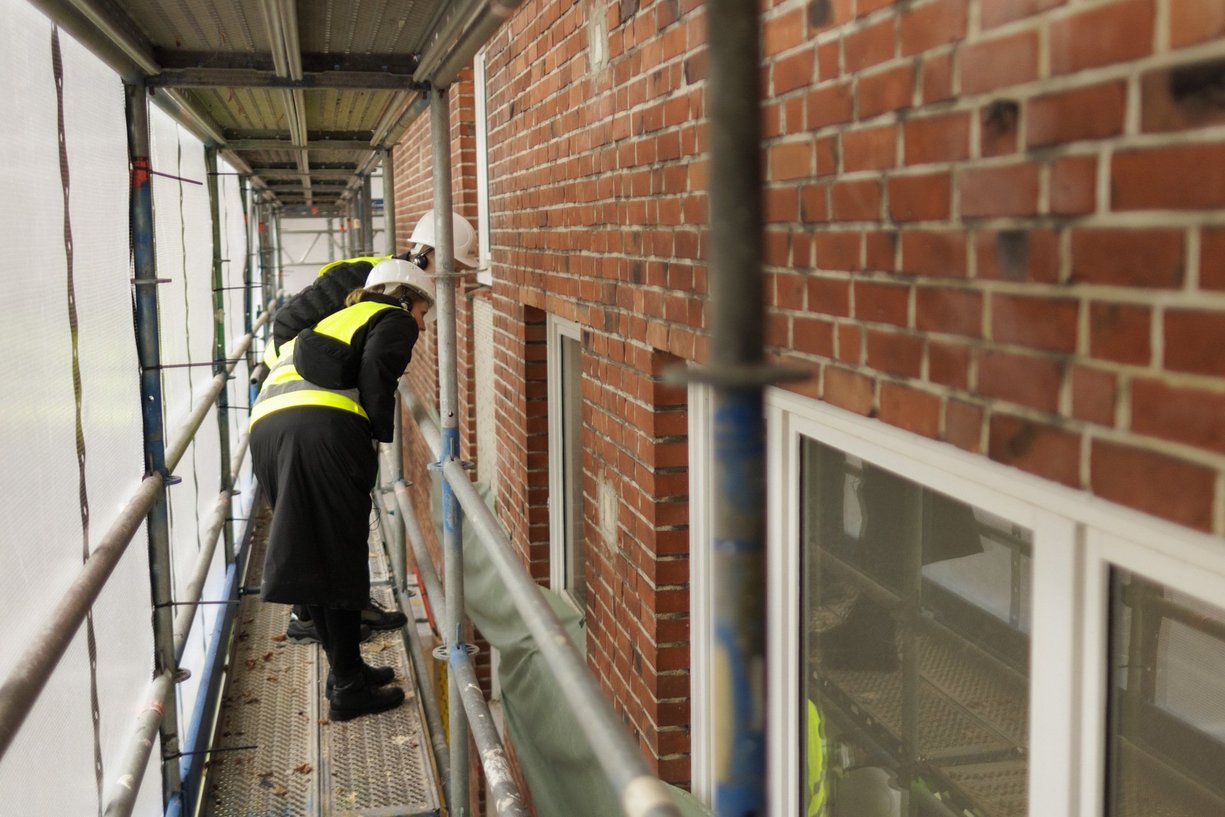Zwei Personen stehen auf einem Baugerüst und schauen in ein Fenster des eingerüsteten Gebäudes.