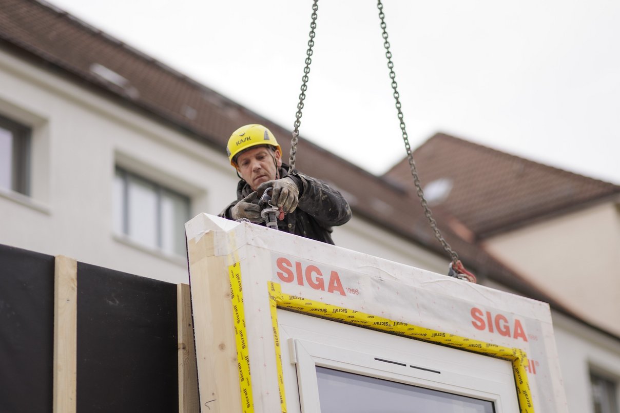 Bauarbeiter mit vorgfertigtem Fassadenelement an Kran, im Hintergrund ein Gebäude