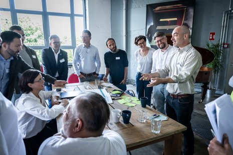 Mehrere Personen stehen und sitzen um einen Tisch herum in einer Workshop-Situation.