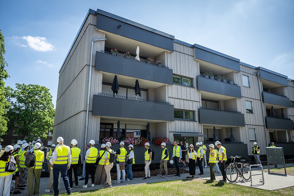 Personen mit gelben Sicherheitswesten stehen vor einem Mehrfamilienhaus.