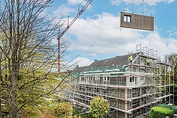 Blick auf ein Mehrfamilienhaus mit Baugerüst. Daneben steht ein Kran, an dem ein vorgefertigtes Fassadenelement schwebt.