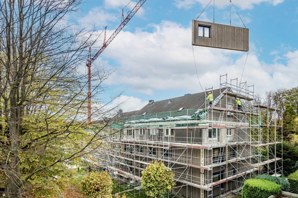 ecoworks wird die Fassaden in Zukunft gemeinsam mit der Oikos Group fliegen lassen. Das erste gemeinsame Projekt wird die serielle Sanierung eines Plattenbaus aus den 70er Jahren in Ost-Berlin sein. Blick auf ein Mehrfamilienhaus mit Baugerüst. Daneben steht ein Kran, an dem ein vorgefertigtes Fassadenelement schwebt.