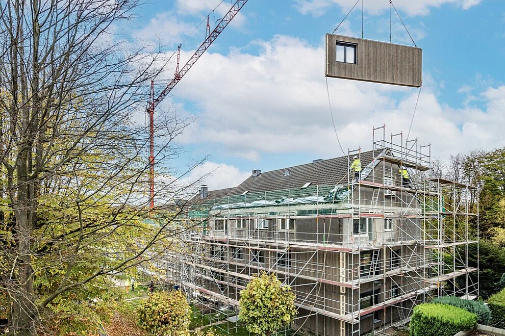 Blick auf ein Mehrfamilienhaus mit Baugerüst. Daneben steht ein Kran, an dem ein vorgefertigtes Fassadenelement schwebt.