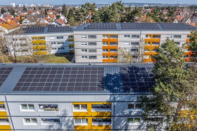 Drei Mehrfamilienhäuser drei- bis viergeschossig mit Photovoltaikanlagen auf den Flachdächern. Im Hintergrund Einfamilienhäuser und Bäume.