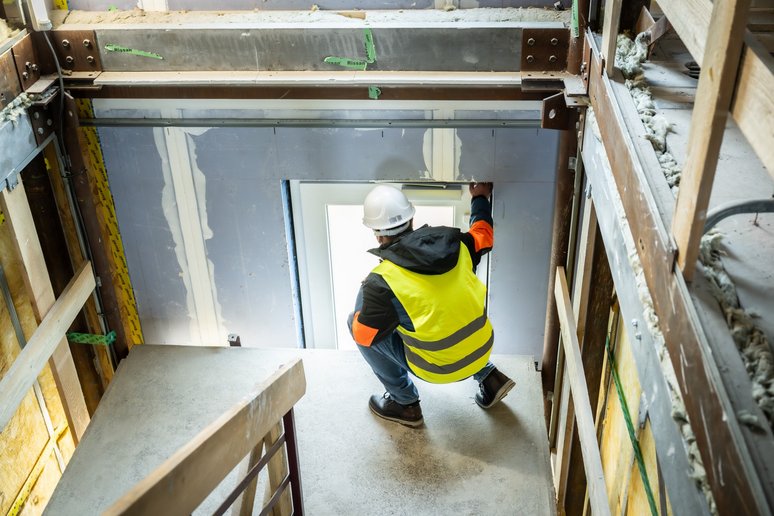 Person mit Bauhelm auf dem Knopf kniet in einem Gebäude am Fenster und schaut hinaus.