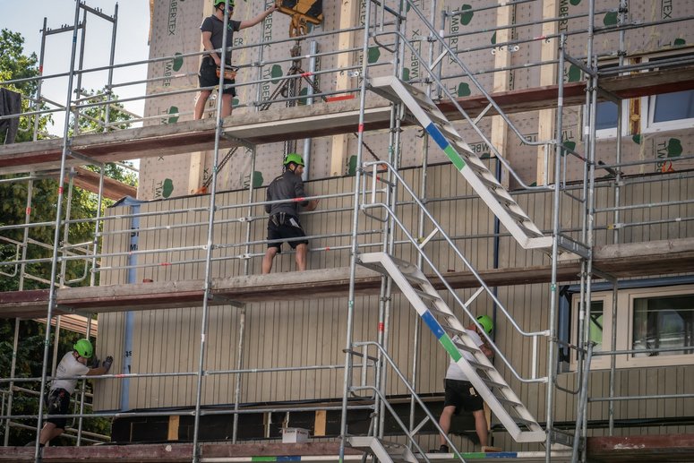 Wohngebäude mit Baugerüst während Sanierung, Bauarbeiter montieren vorgefertigte Fassadenelemente.