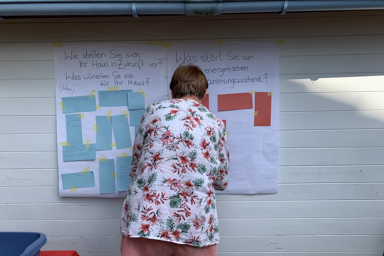 „Wie wünsche ich mir mein Haus in 10 Jahren?“ – Zukunftsvisionen für das eigene Zuhause. Frau steht mit dem Rücken vor Plakaten und Klebezetteln.