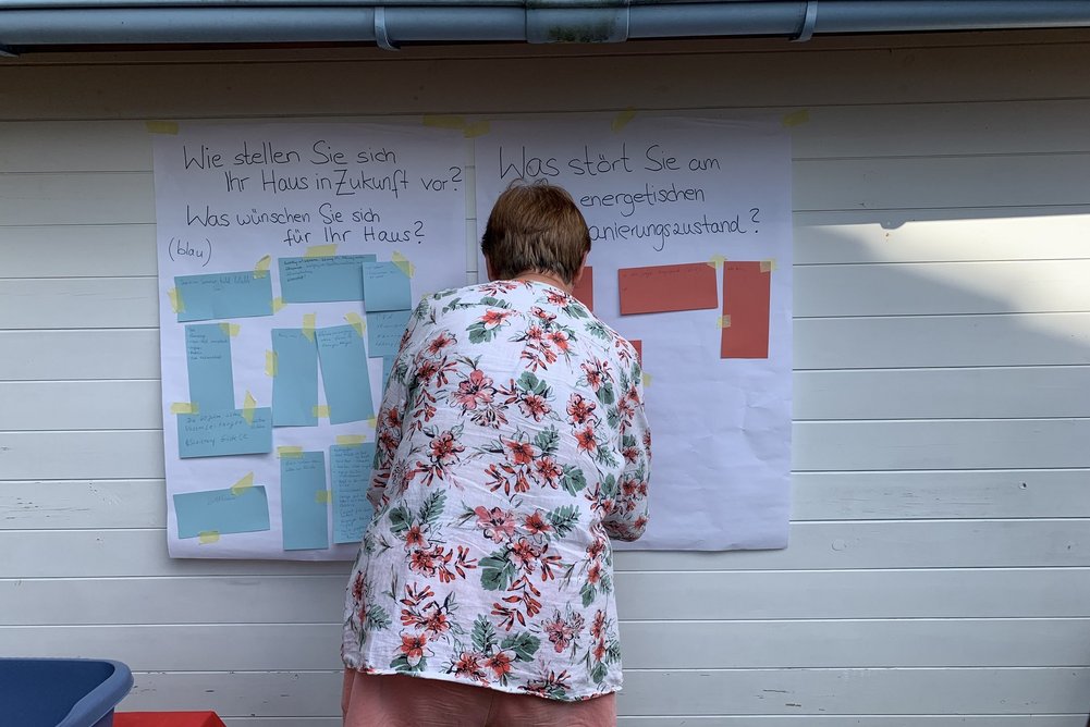 „Wie wünsche ich mir mein Haus in 10 Jahren?“ – Zukunftsvisionen für das eigene Zuhause. Frau steht mit dem Rücken vor Plakaten und Klebezetteln.