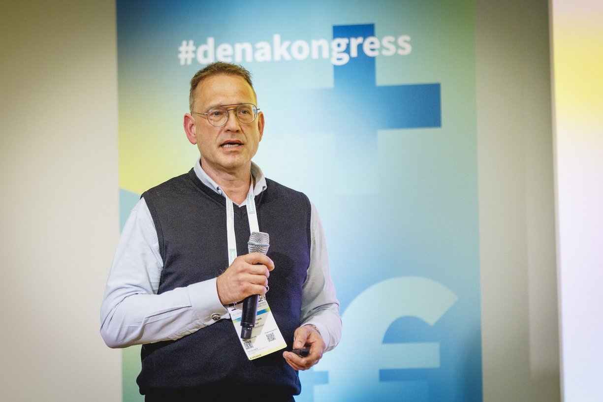 Prof. Dr. Frank Wolter, Leiter Kompetenzteam Exzellenz & Innovation der DB-Immobilien Sprechende Person mit Mikrofon in der Hand, Hintergrundbild zum dena Kongress.