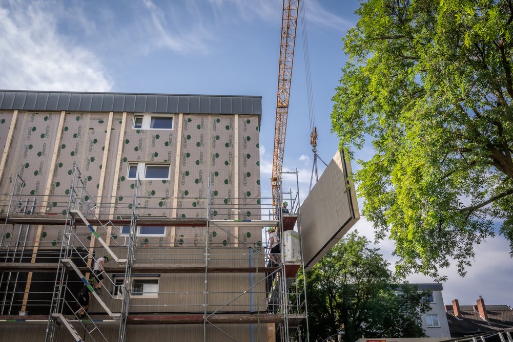 Bild zeigt: Kran mit hängendem Fassadenelement neben Gebäude im Baugerüst