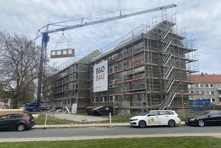 Kran mit vorgefertigtem Fassadenelement, dass über einem Mehrfamilienhaus schwebt. Davor ist eine Straße mit parkenden PKWs.