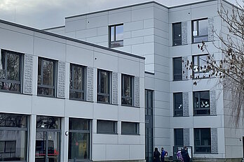 Schulgebäude mit heller Fassade, im Hintergrund bewölkter Himmel