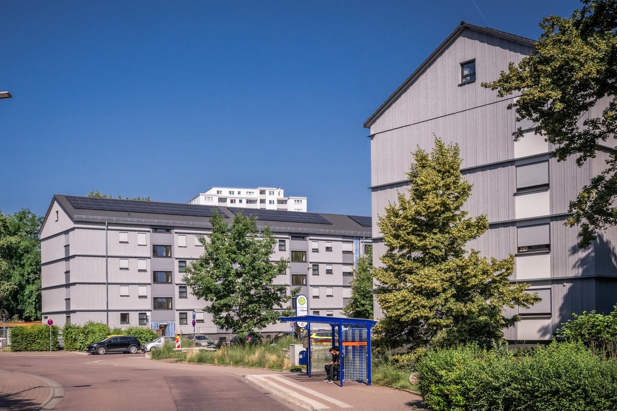 Straße durch ein Wohngebiet, auf der rechten Seite zwei neu sanierte Mehrfamilienhäuser viergeschossig und Grünanlagen