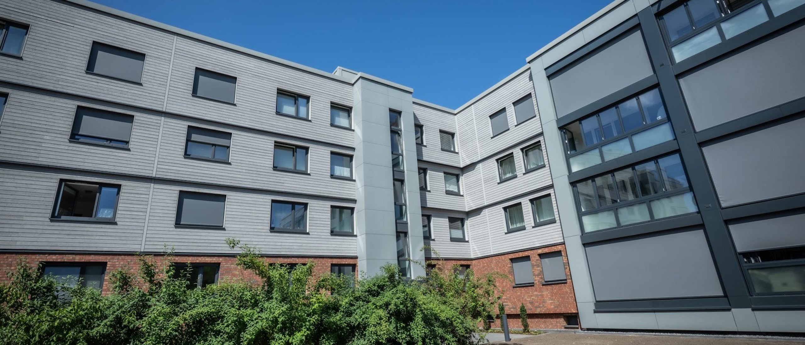 Mehrfamilienhaus viergeschossig nach Sanierung, davor Grünanlagen, im Hintergrund blauer wolkenloser Himmel