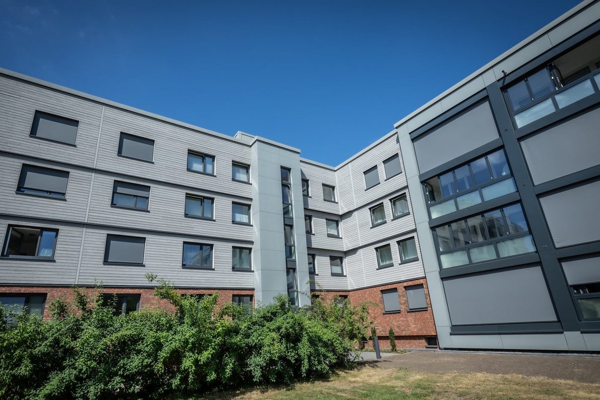 Mehrfamilienhaus viergeschossig nach Sanierung, davor Grünanlagen, im Hintergrund blauer wolkenloser Himmel