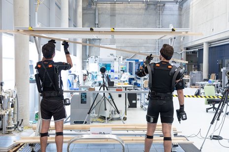 Exoworkathlon, zwei Personen (rückseitig) in spezieller Arbeitskleidung, im Hintergrund eine Halle mit Arbeitsplätzen und technischer Ausstattung
