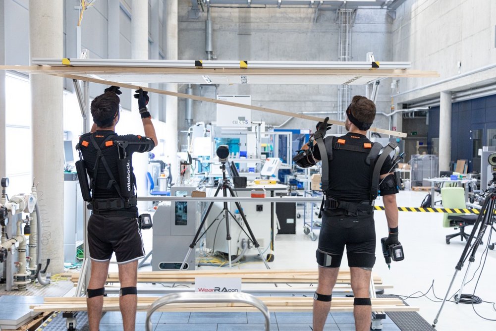 Exoworkathlon, zwei Personen (rückseitig) in spezieller Arbeitskleidung, im Hintergrund eine Halle mit Arbeitsplätzen und technischer Ausstattung