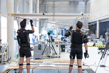 Exoworkathlon, zwei Personen (rückseitig) in spezieller Arbeitskleidung, im Hintergrund eine Halle mit Arbeitsplätzen und technischer Ausstattung