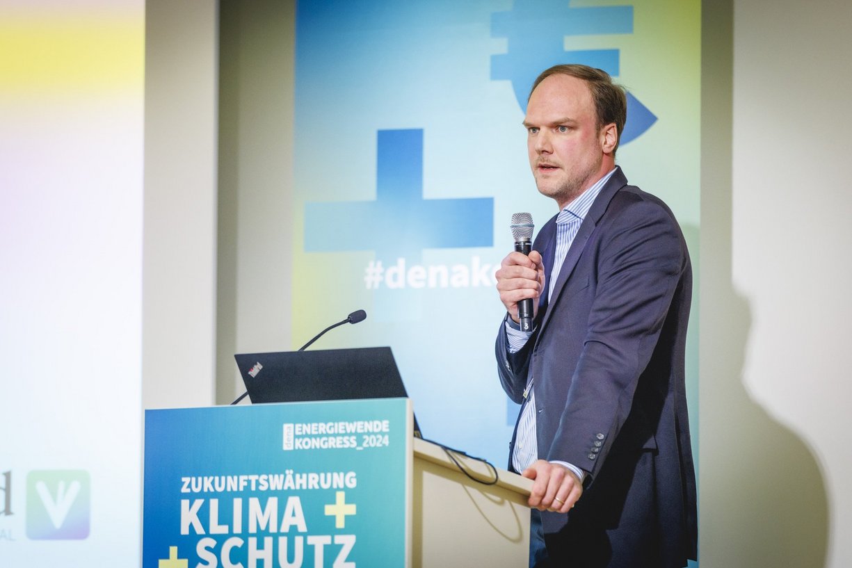 Axel Tønnesen, Head of Business Development der vreed GmbH Person im Anzug mit Mikrofon in der Hand steht am Renderpult und spricht.