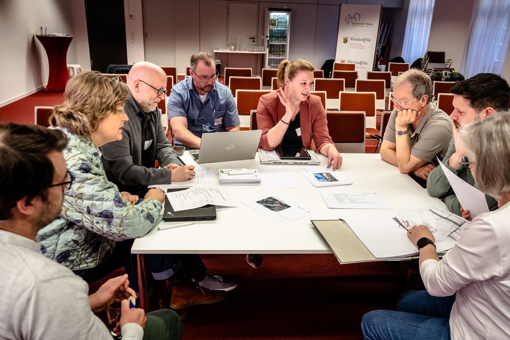 Acht Personen sitzen an einem großen Tisch in einem Veranstaltungsraum. Auf dem Tisch sind Notebooks und weitere Materialien.