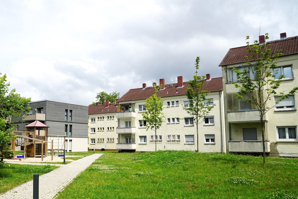 Die hellen Häuser gehören zu denen, die seriell energetisch saniert und deren Dächer aufgestockt werden. Die neue Fassade wird dem ebenfalls von B&O gebauten Holzhybridhaus hinter dem Spielplatz angeglichen. Mehrfamilienhäuser dreigeschossig mit Schrägdach, Grünanlagen und Spielplatz