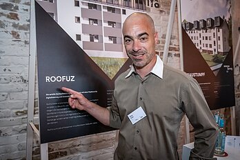 Mann vor Plakat mit saniertem Haus und Aufschrift "Roof Uz"