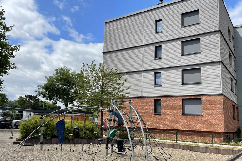 Seitlicher Blick auf ein viergeschossiges Wohngebäude mit Flachdach, davor ein Kinderspielplatz