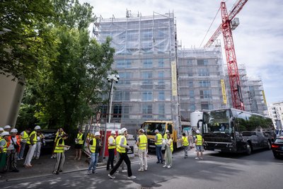 Großer Gebäudekomplex während Sanierung, davor ein Baukran und ein parkender Reisebus. Personen mit gelben Sicherheitswesten laufen auf einem Fußweg.