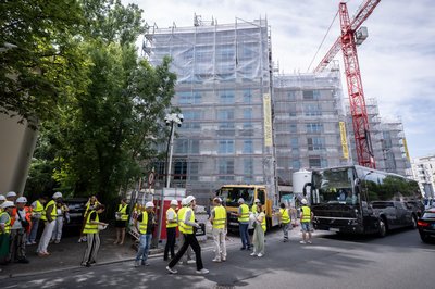 Großer Gebäudekomplex während Sanierung, davor ein Baukran und ein parkender Reisebus. Personen mit gelben Sicherheitswesten laufen auf einem Fußweg.