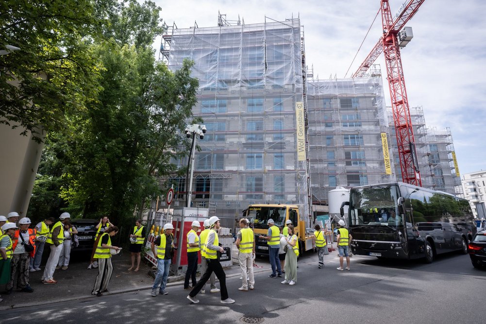 Großer Gebäudekomplex während Sanierung, davor ein Baukran und ein parkender Reisebus. Personen mit gelben Sicherheitswesten laufen auf einem Fußweg.