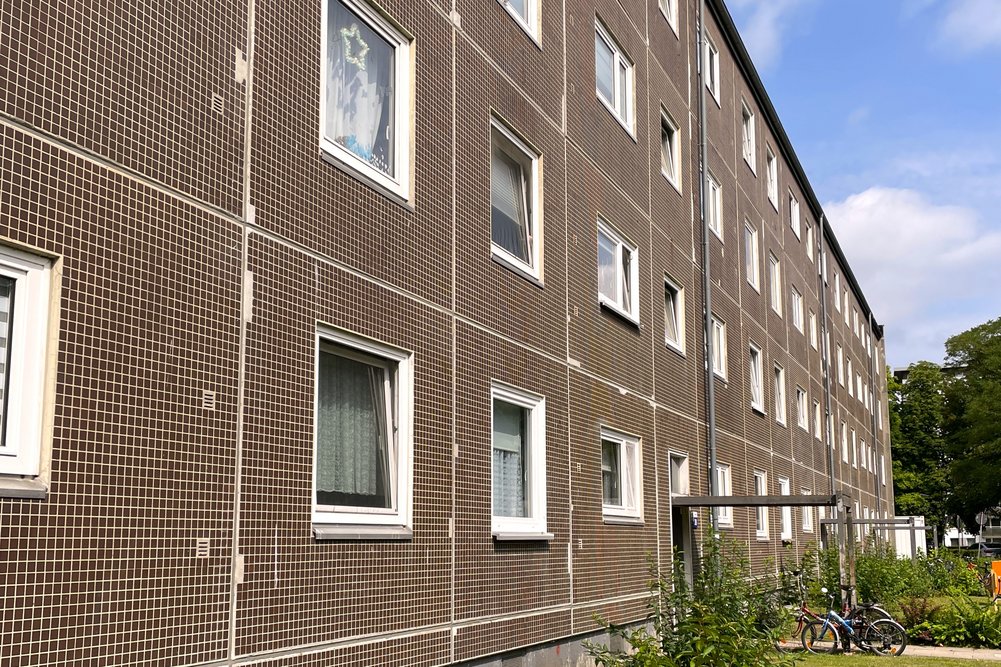 Vor der Sanierung Wohngebäude mit dunkler Ziegelfassade und weißen Fenstern, umgeben von einem Rasenbereich mit Sträuchern und Fahrrädern unter leicht bewölktem Himmel.