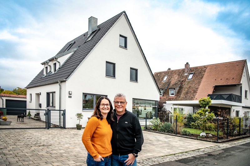 Zwei Personen stehen auf einem Weg, im Hintergrund ein saniertes Eigenheim mit weißer Putzfassade und Schrägdach