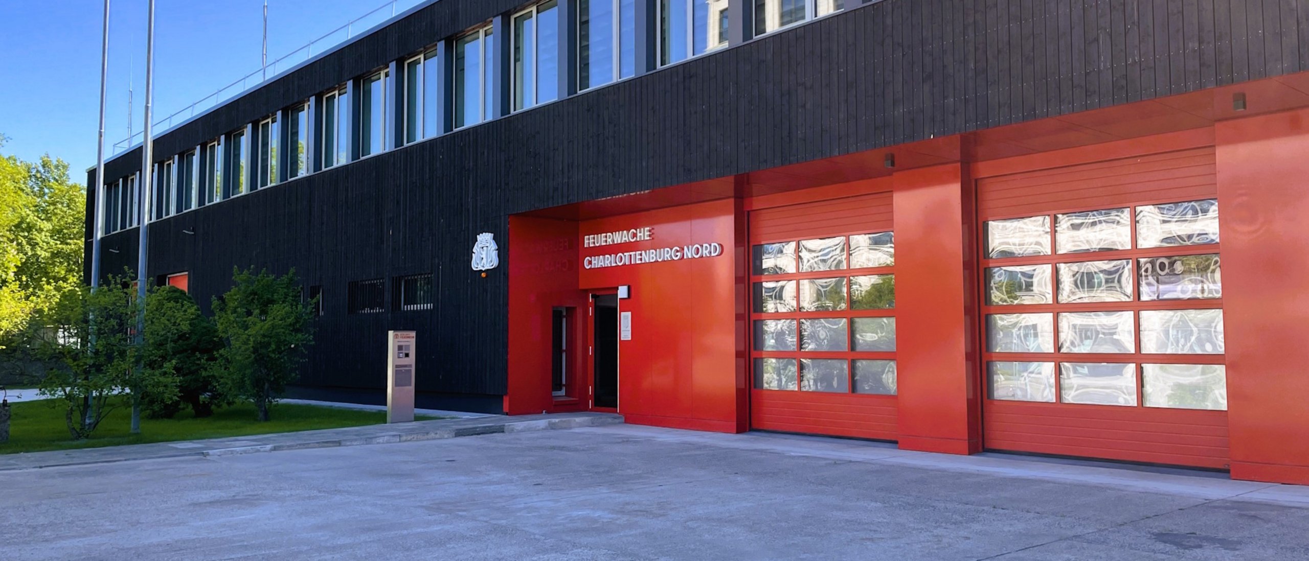 Außenansicht Feuerwache Charlottenburg nach Sanierung, Gebäude zweigeschossig, dunkle Holzfassade mit Fenstern, rote Torzufahrt