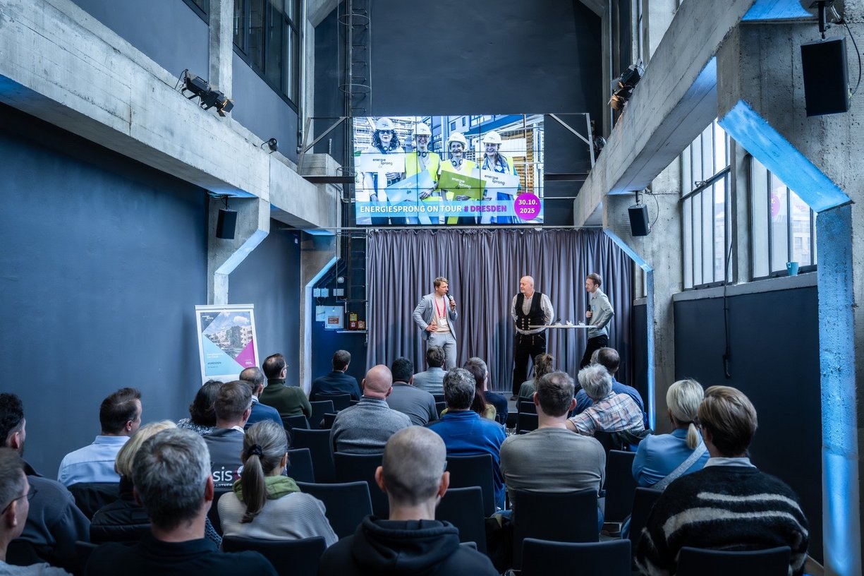 Personen sitzen auf Stühlen in einem Veranstaltungsraum und schauen zur Bühne mit Vortragenden.
