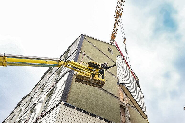 Haus mit teilweise neuer Fassade und Arbeiter der von einem Kran aus vorgefertigte Elemente entgegen nimmt.