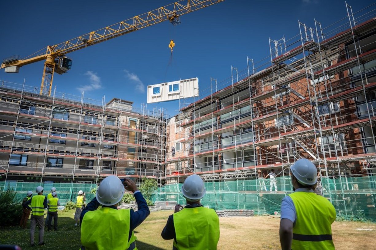 Personen mit gelben Sicherheitswesten und Bauhelmen schauen auf ein Mehrfmailienhaus. An dem Haus steht ein Baugerüst. An einem Kran schwebt ein vorgefertigtes Hausfassadenelement.