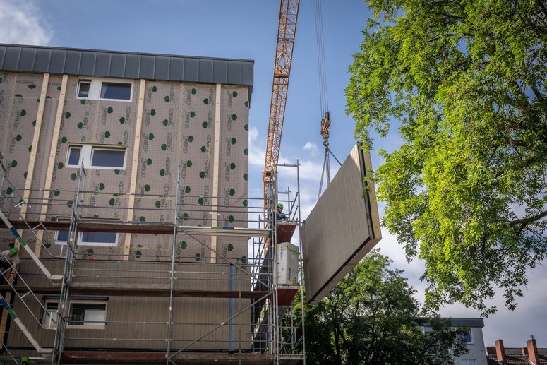 Wohngebäude während der Sanierung, Kran mit einem vorgefertigten Fassadenelement, im Vordergrund ein Laubbaum