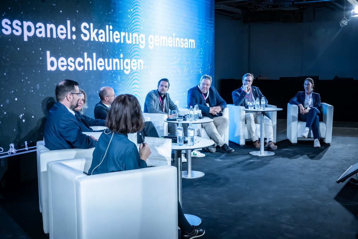 Bühne mit weißen Sitzmöbeln, Panel-Talk mit acht Personen bei einer Fachveranstaltung