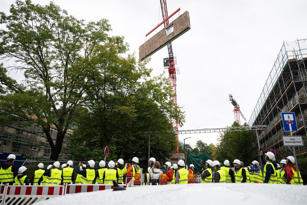 Heinrich-Hertz-Quartier in Erlangen-Bruck Mehrere Personen in gelber Warnweste und Helm auf dem kopf auf einer Baustelle, schwebendes Fassadenelement