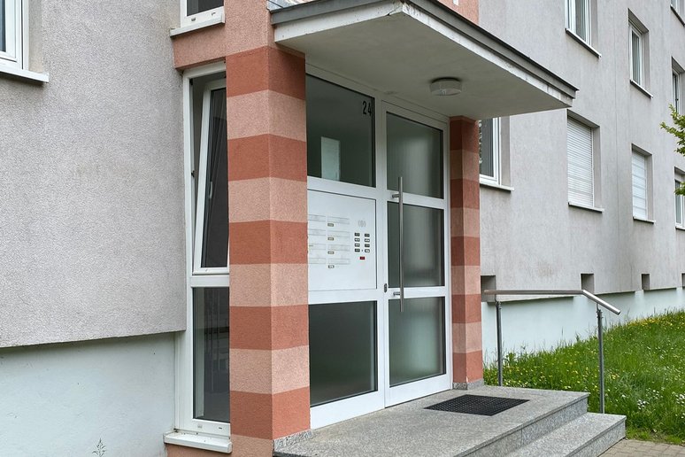 Nahaufnahme des Eingangs eines Mehrfamilienhauses mit der Hausnummer 24. Der Eingang liegt unter einem Vordach und ist über mehrere Stufen mit Handlauf erreichbar. Die Fassade ist hellgrau verputzt und wird von zwei rosa- und terrakottafarben gestreiften Pfeilern eingefasst. Neben dem Eingang befinden sich Fenster und eine Rasenfläche.
