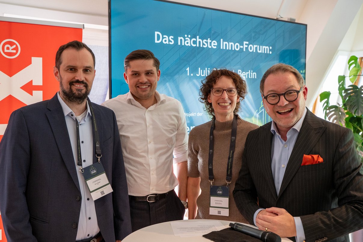 Vier Personen stehen an einem Tisch nebeneinander für ein Foto, im Hintergrund eine Präsentationsfläche mit Text: Das nächste Inno-Forum