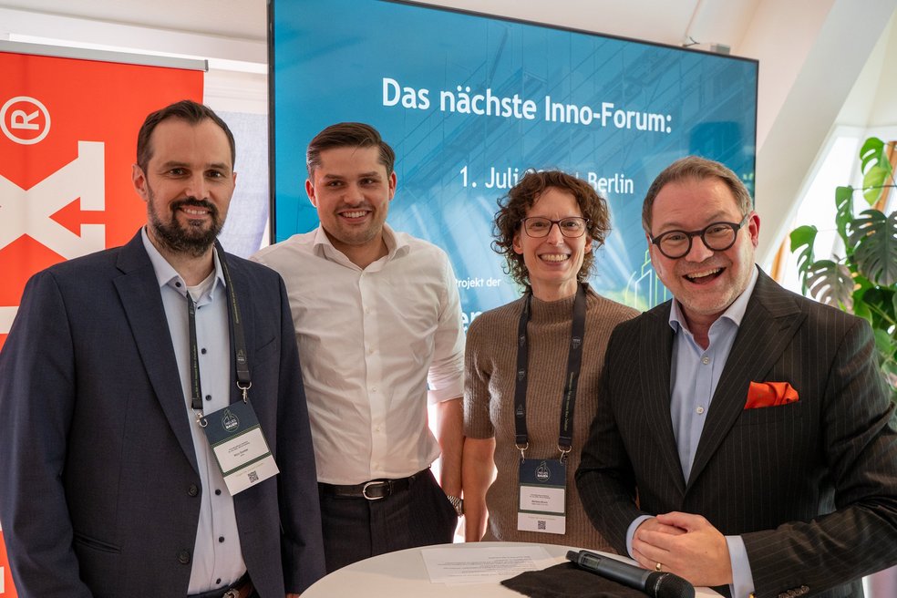 Vier Personen stehen an einem Tisch nebeneinander für ein Foto, im Hintergrund eine Präsentationsfläche mit Text: Das nächste Inno-Forum