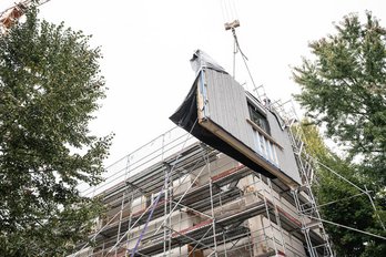 Vorgefertigtes Fassadenelement schwebt an Kran vor Mehrfamilinehaus mit Baugerüst, links und rechts sind Laubbäume.