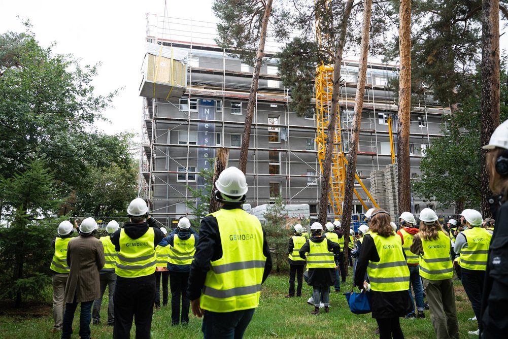 Quartier Jaminstraße Mehrere Personen mit Warnweste und Helm auf dem Kop auf einer Baustelle blicken zum schwebenden Bauelemente.